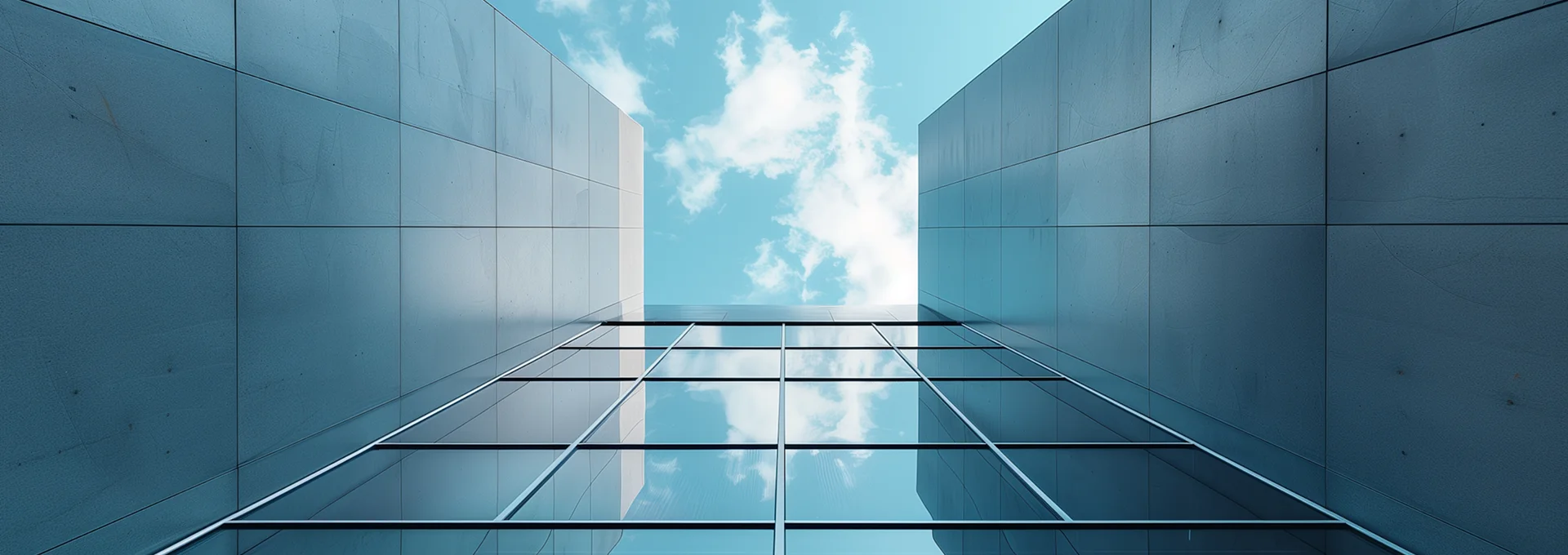 Looking up between two modern glass and steel buildings with a blue sky and clouds above.